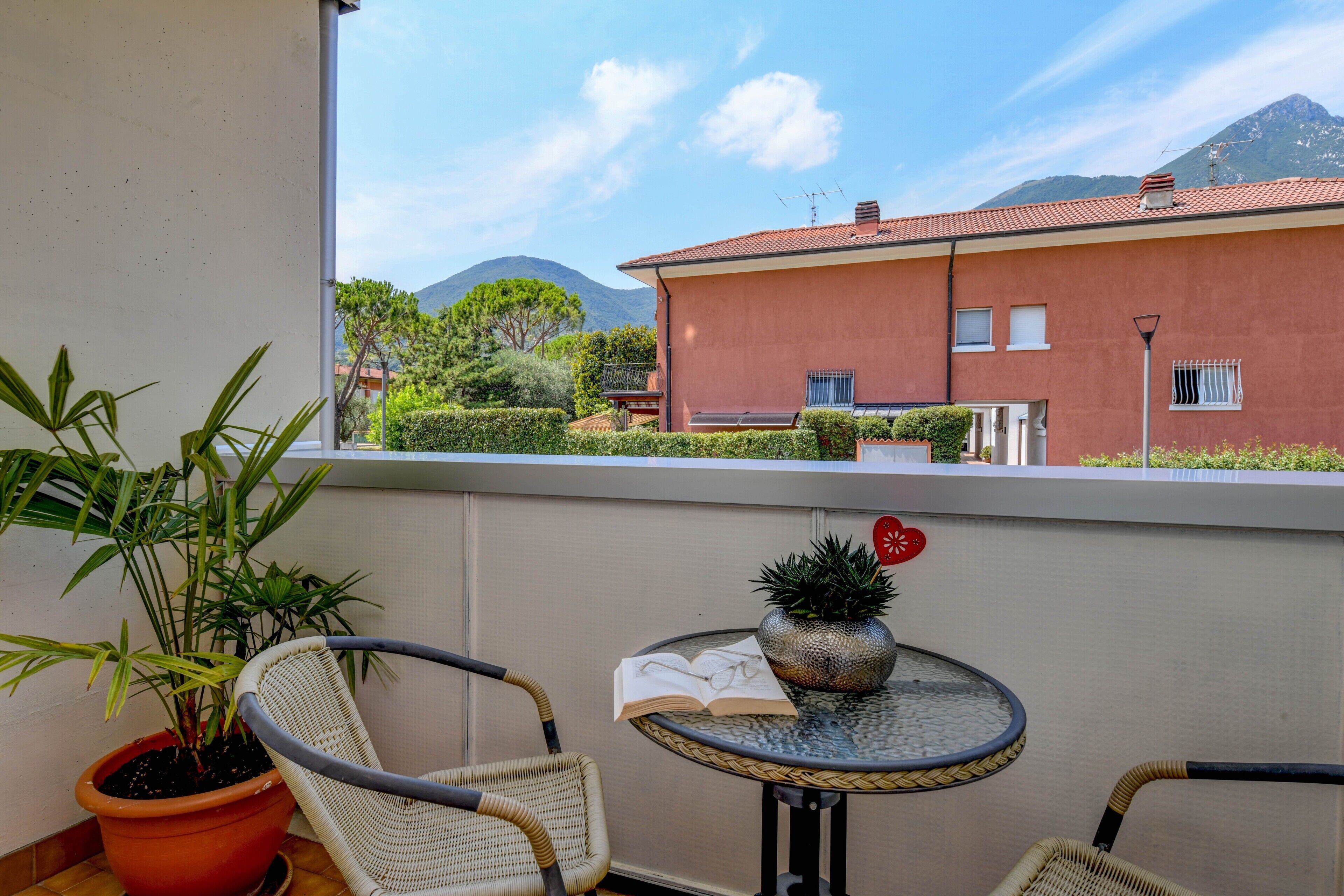 A balcony with wicker chairs, a small table with a plant and an open book, and a view of a red building with a mountain in the background.