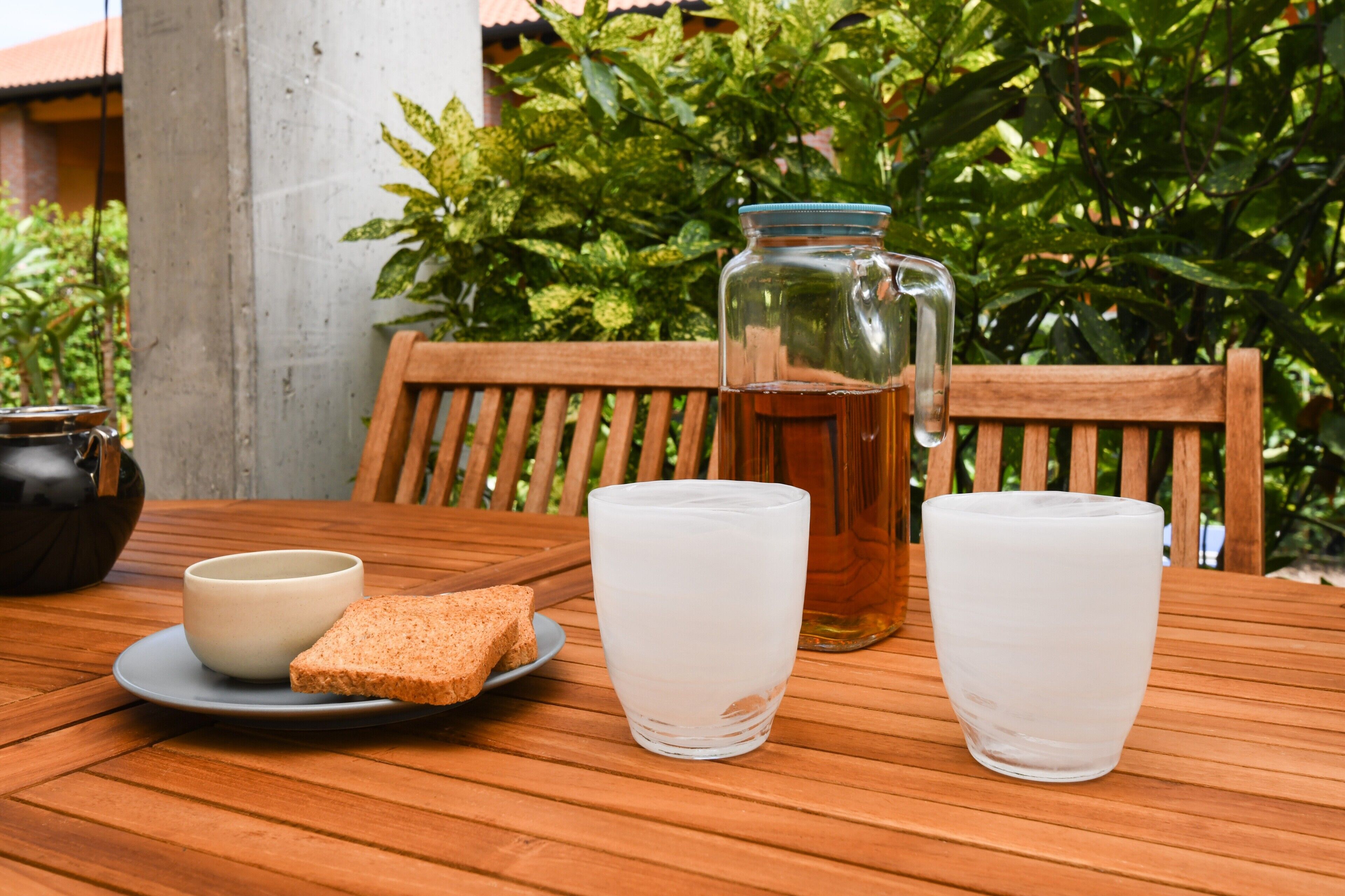 Uma mesa de madeira com um copo de água, um copo de leite, um jarro de chá e um prato com duas fatias de pão.