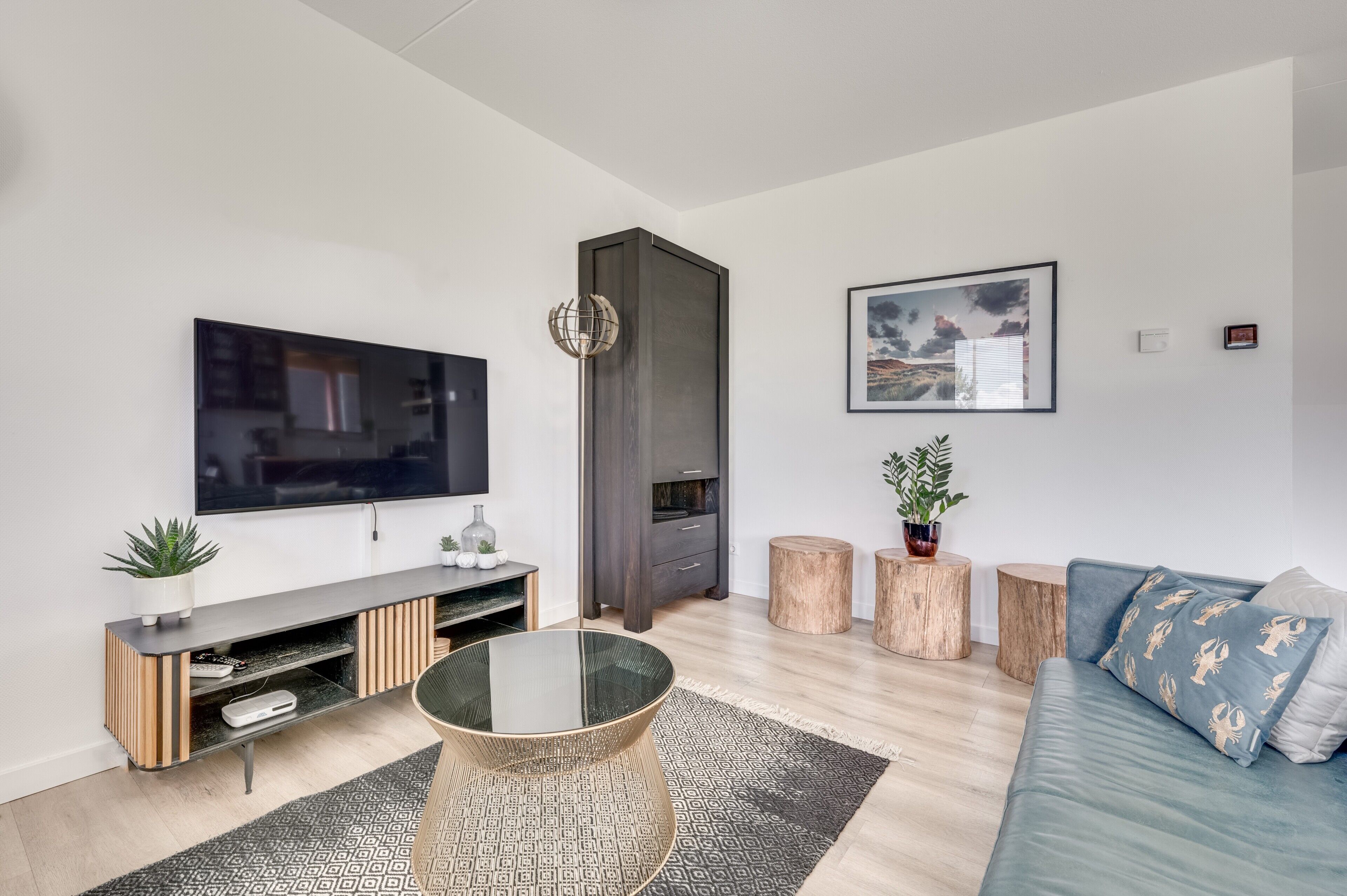 A modern living room with a flat-screen TV, a dark entertainment unit, a glass coffee table, a blue sofa, and a framed landscape painting.