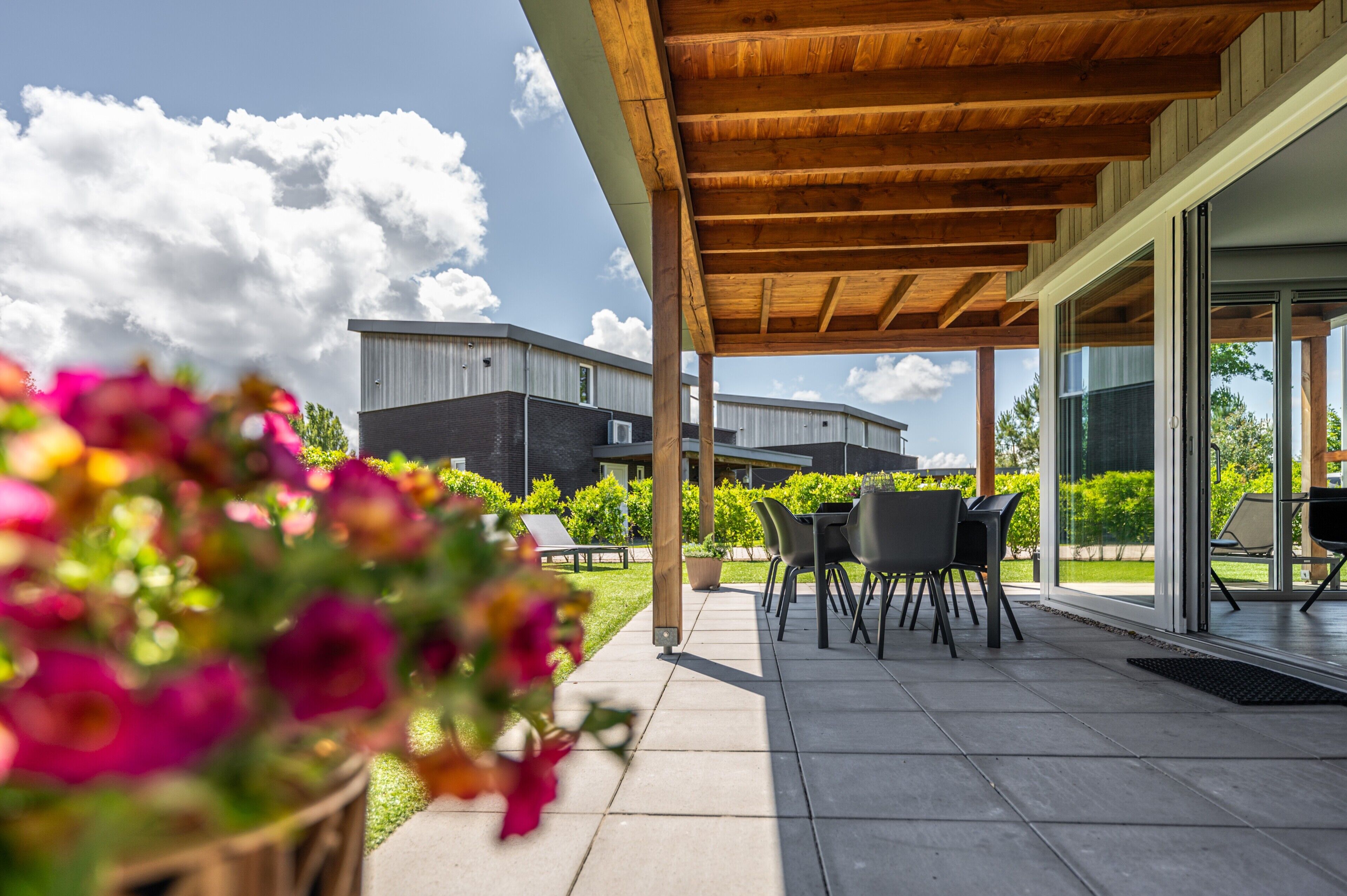 Een overdekt terras met buitenmeubilair, uitzicht op de gevel van het gebouw, en een tuin met bloemen.