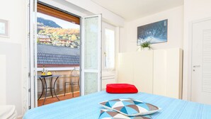 A bedroom with a bed, a red pillow, a window with a view of houses and trees, and a door. - Mountain Views in the Historic Center - Liver 201 (Pieve di Tremosine sul Garda BS)