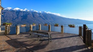 A bench on a paved terrace with a view of snow-capped mountains and a body of water. - Mountain Views in the Historic Center - Liver 201 (Pieve di Tremosine sul Garda BS)