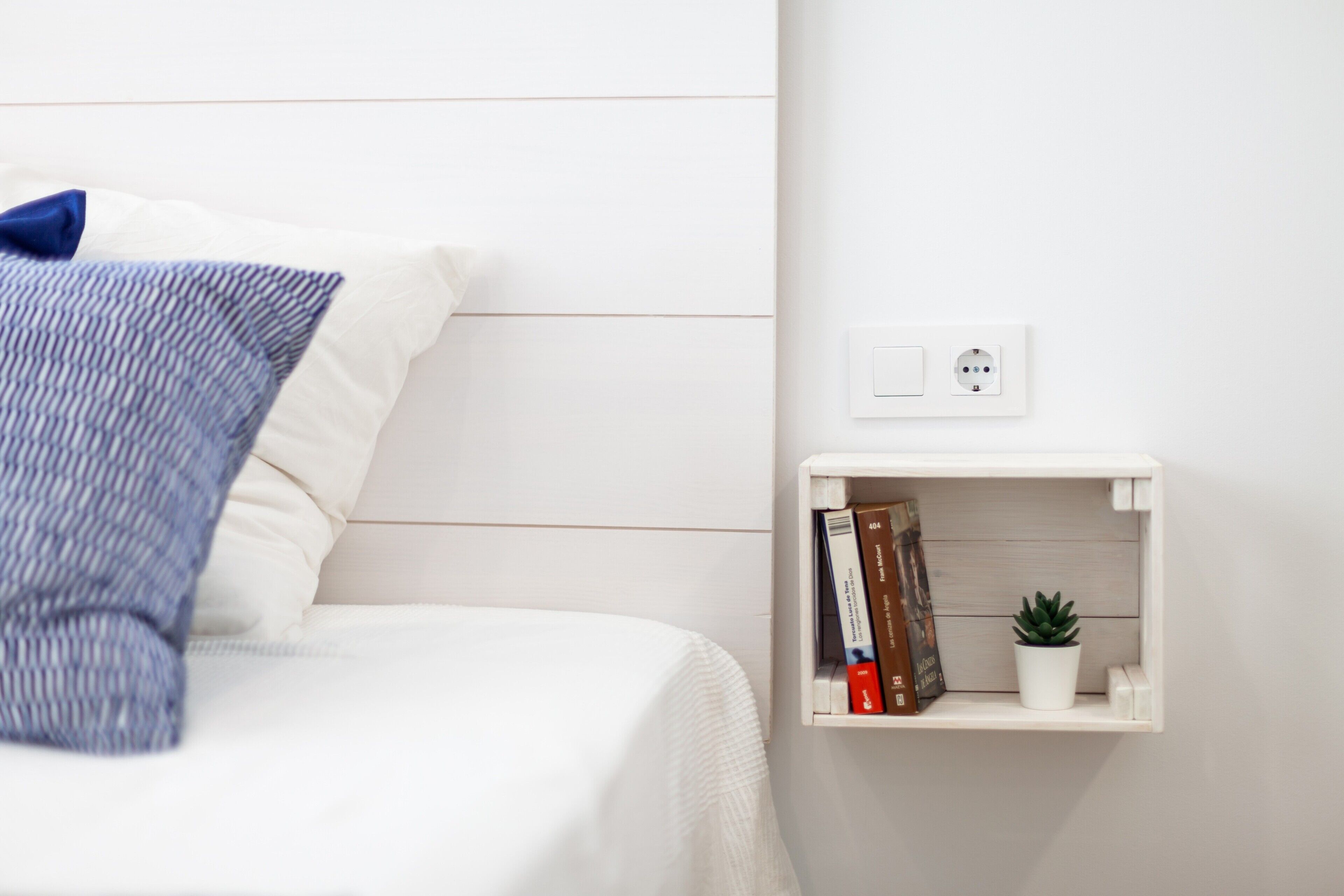 A neatly made bed with a blue pillow, a white bedside table with a small plant, and a power outlet.