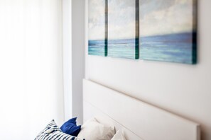 Une chambre avec une tête de lit blanche, un lit recouvert d’une couette à rayures bleues et blanches, et un tableau représentant un paysage marin.