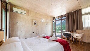A hotel room with a wooden ceiling, a large bed with white linens, a red throw blanket, a desk with a chair, and a view of a mountainous landscape through the window. - With panoramic views in the hills – Apartment GeoResort A2 (Tremosine sul Garda)