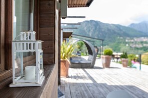 Una terraza de madera con un farol blanco, una planta en maceta y vistas a montañas.