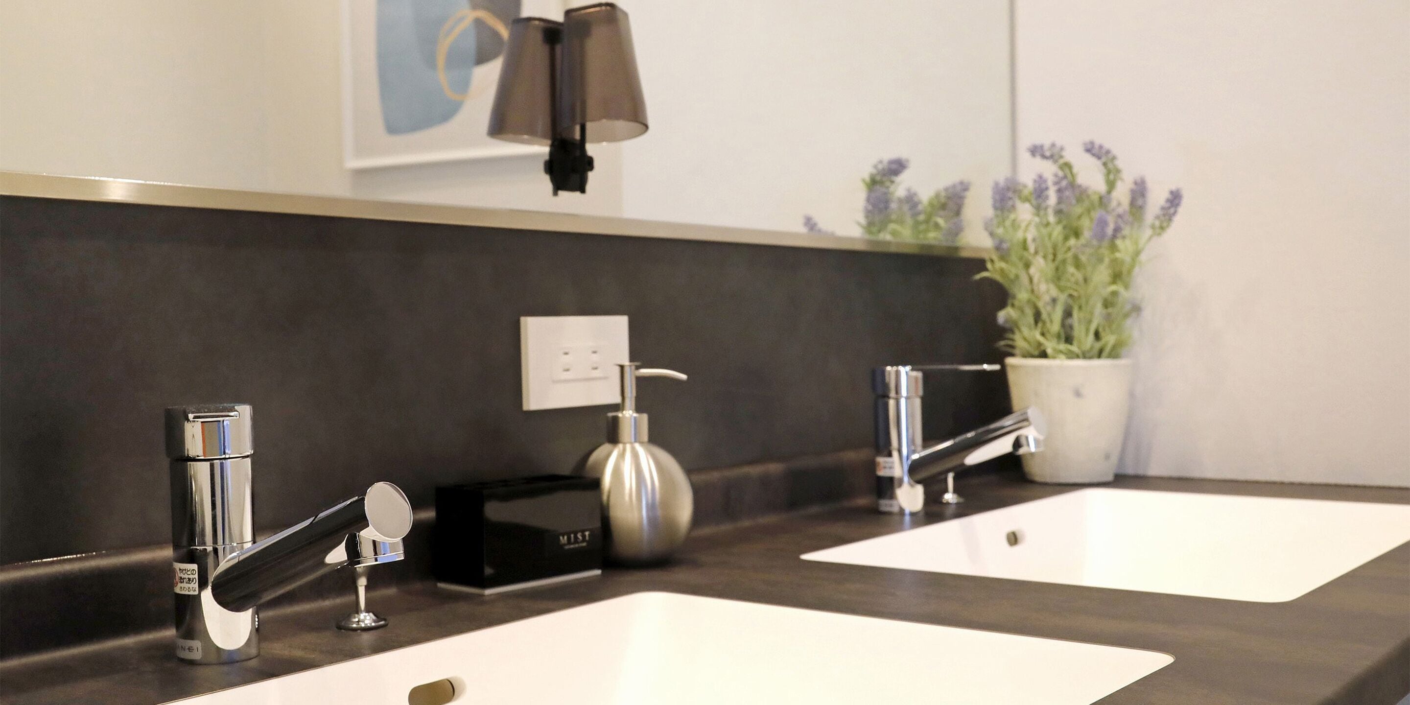 A modern bathroom with two sinks, a mirror, a wall-mounted light, and a potted lavender plant.
