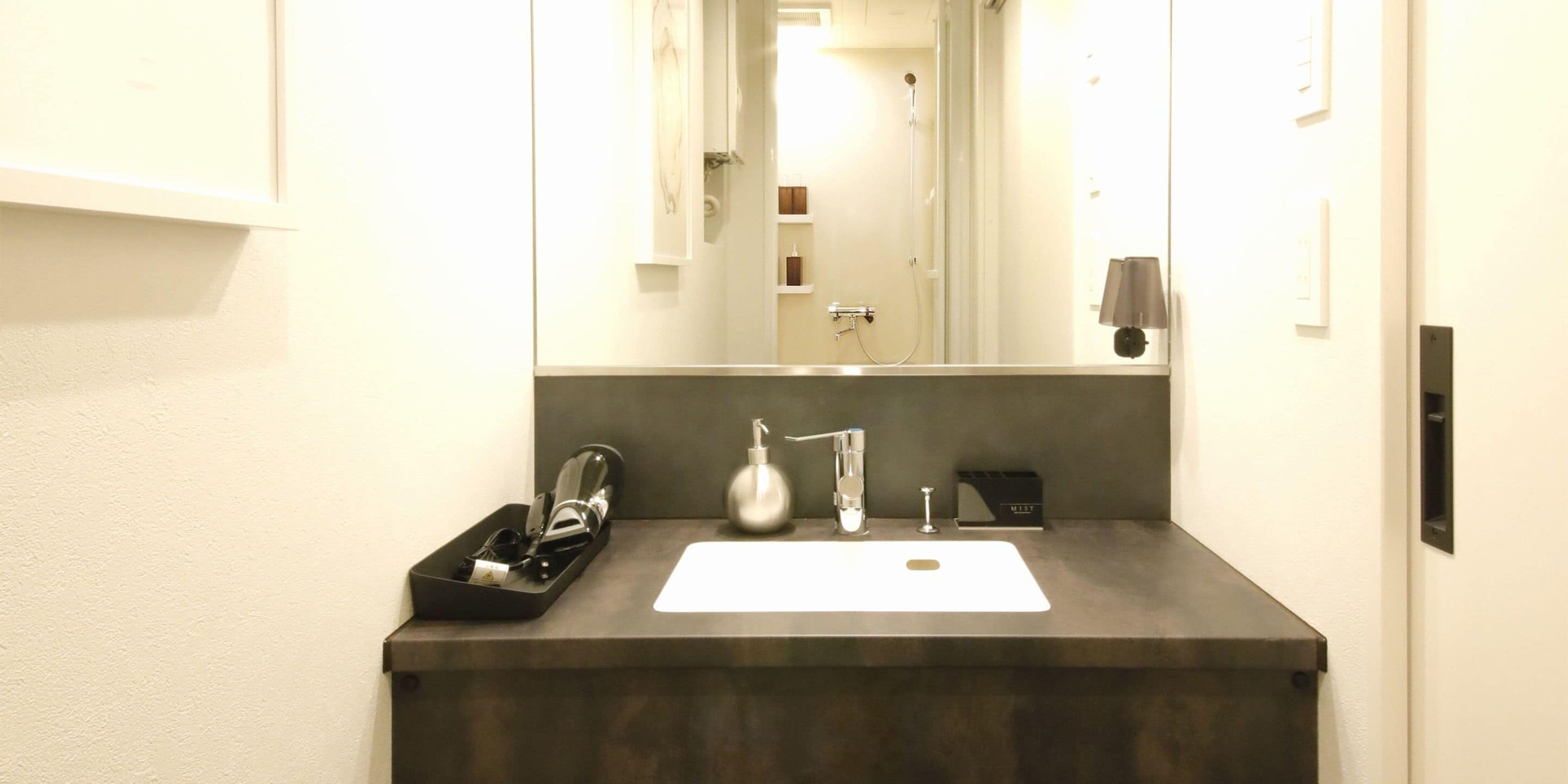 Un bagno moderno con un piano d'appoggio scuro, un lavandino bianco, uno specchio e una doccia visibile nel riflesso.
