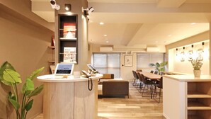A reception area with a wooden desk, a laptop, and a potted plant. There is a dining area with a table and chairs in the background. - Lestel Maru - Hostel (Sapporo)