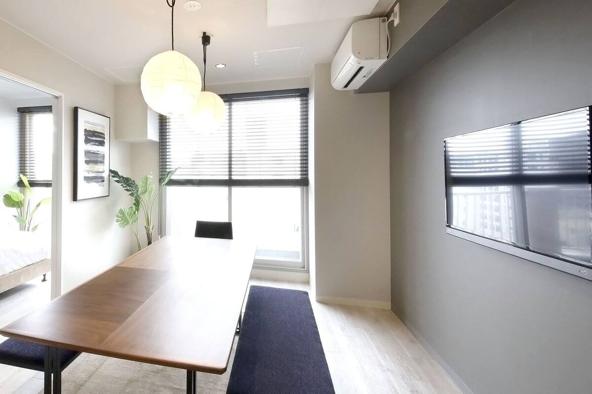 A modern, well-lit room with a long wooden table, a bench, and a large window with blinds.