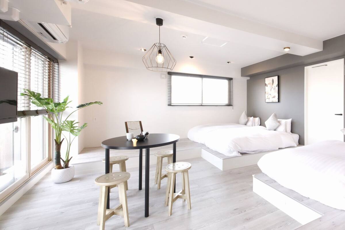 A modern bedroom with a large bed, a smaller bed, a dining area with stools, and a potted plant.