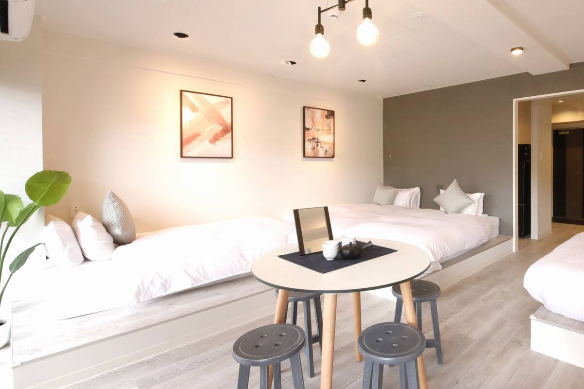A modern hotel room with a large bed, a smaller bed, a round dining table with stools, and artwork on the walls.