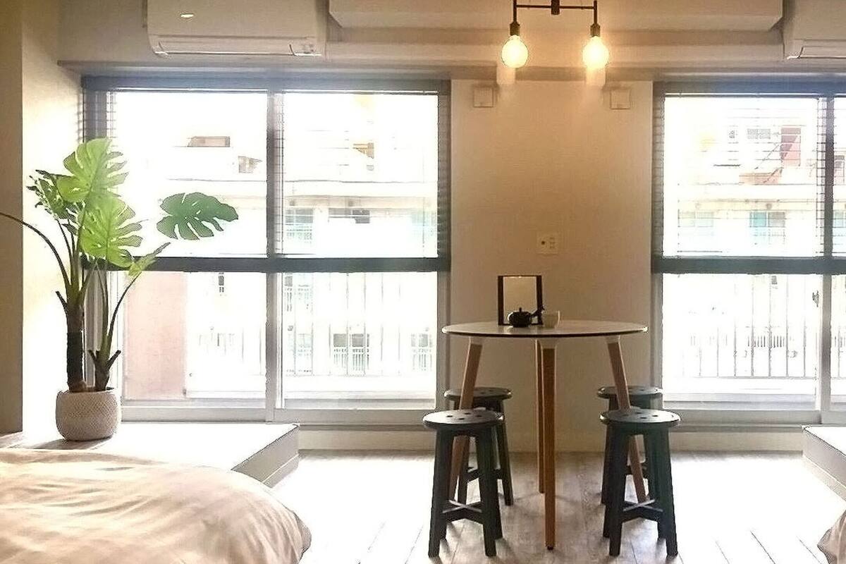 A room with two beds, a small table, two stools, and large windows with a view of buildings.
