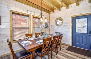 Un coin repas avec une table en bois dressée pour six personnes, entourée de chaises en bois. Un lustre est suspendu au-dessus de la table, et une porte bleue se trouve à droite.