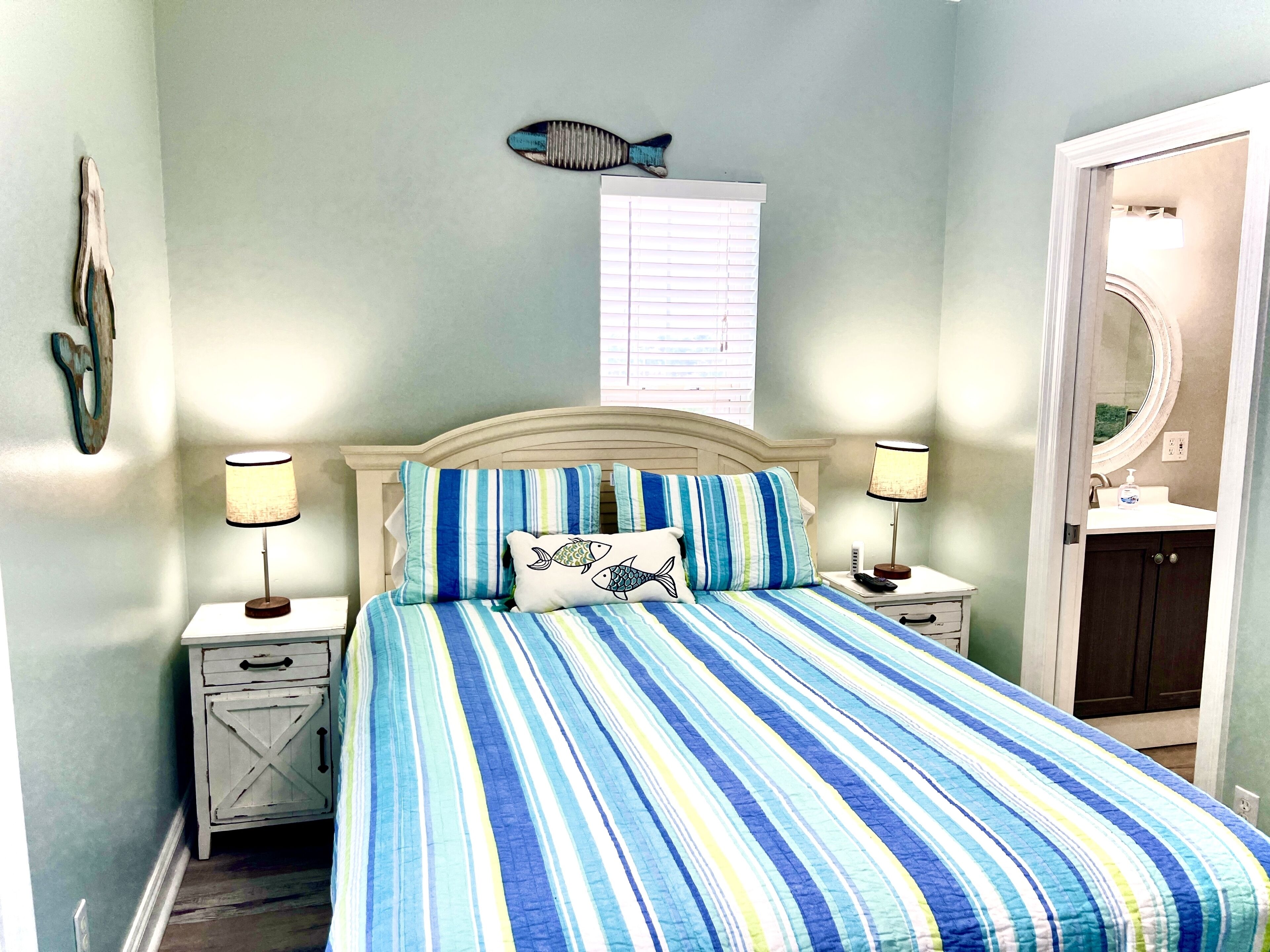 A bedroom with a striped bedspread, two bedside lamps, a fish decoration on the wall, and a nightstand with a drawer.