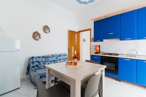 Ett kompakt kök med blå skåp, ett matbord med stolar, ett kylskåp och en liten köksdel.