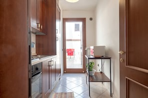 A narrow hallway with a kitchen area on the left, a small table with a plant on the right, and a door leading to an outdoor area.