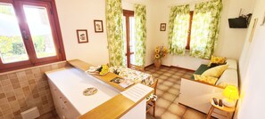 A kitchen with a dining table, chairs, a sofa, a small table with a lamp, and a TV mounted on the wall. - With a terrace and garden immersed in nature - Apartment Wanna B (Capoliveri (LI))