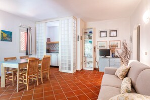 Espaço de refeições com mesa e cadeiras, cozinha com armários e televisão, e sala de estar com sofá e objetos decorativos.