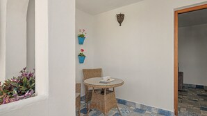Ein kleiner Sitzbereich im Freien mit einem runden Tisch und Korbstühlen, zwei Kübelpflanzen an der Wand und einem Fliesenboden mit Muster.