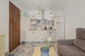A compact kitchen with white cabinets, a microwave, and a coffee maker. There is a wooden door and a brown sofa in the living area.