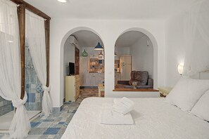 A bedroom with a large bed, an arched doorway leading to a bathroom, and a view of a kitchen area.