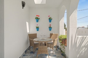 A small outdoor seating area with a round table and wicker chairs, surrounded by white walls and blue patterned tiles.