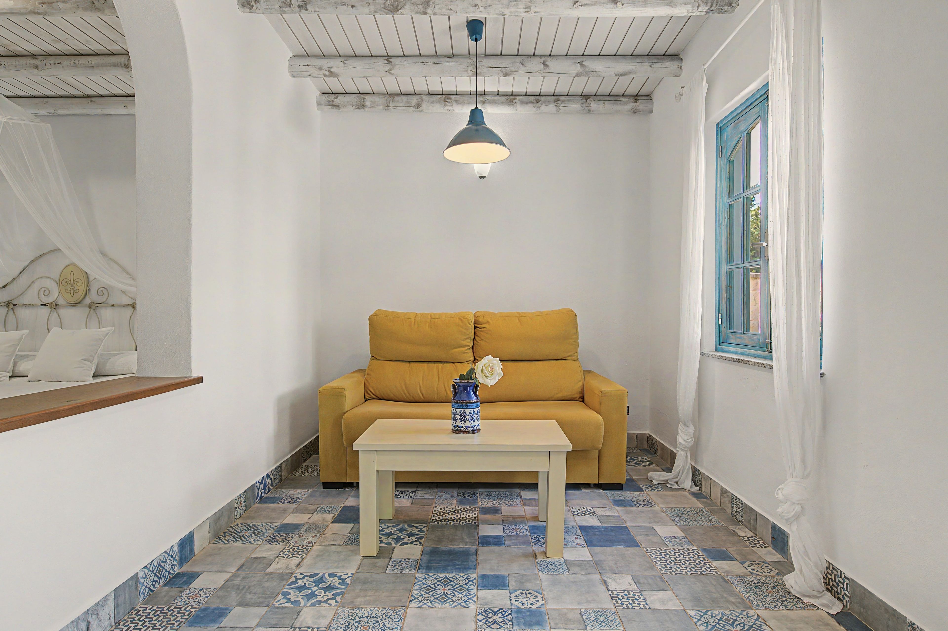 Una habitación con un sofá amarillo, una mesa blanca y un jarrón azul con una flor blanca.