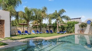 A swimming pool with clear water, surrounded by lounge chairs and umbrellas, with palm trees and a building in the background. - Charming Apartment with Pool, Terrace, Air Conditioning and Wi-Fi (Conil)
