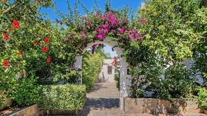 An outdoor pathway leading through a garden with a white wall and an archway covered in flowering plants. - Charming Apartment with Pool, Terrace, Air Conditioning and Wi-Fi (Conil)