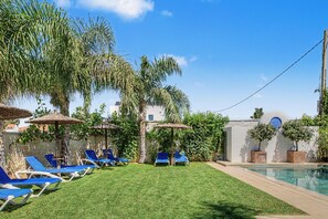 Un espace aménagé au bord de la piscine, avec des chaises longues, une pelouse verdoyante et des palmiers.