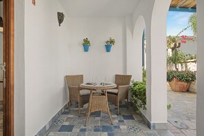 Un petit espace patio avec une table ronde et deux chaises en osier, deux plantes en pot et un sol carrelé.