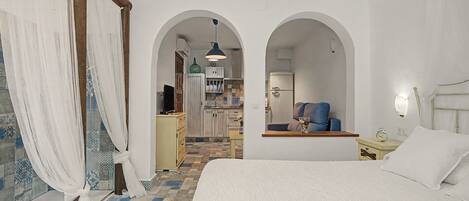A bedroom with a large bed, a nightstand, and a chair. There is a kitchen area visible through an archway.