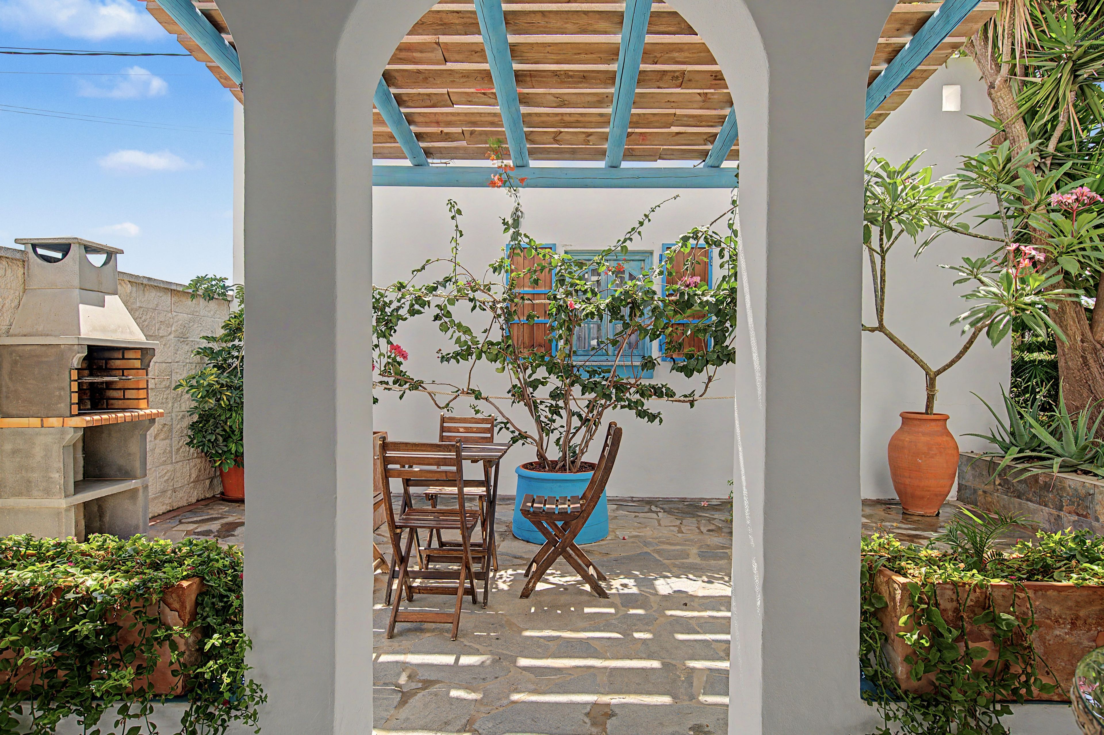 A covered patio area with a wooden table and chairs, a potted plant, and a stone wall.
