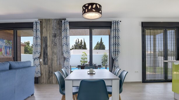 A modern dining area with a table and chairs, a sofa, and a view through large windows. - Modern Holiday Home ''Casa Toria'' with Private Pool, Air Conditioning (Conil de la Fontera)
