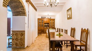 Un comedor con una larga mesa de madera y sillas, una araña y una cocina al fondo.