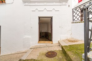 Une étroite entrée mène à un bâtiment à la façade blanche, doté d’un portail en métal et d’un petit seuil qui donne sur une cour intérieure agrémentée de plantes en pot.