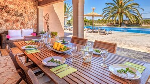 Un espace de restauration en plein air agrémenté d’un mur en pierre, de mobilier en bois et offrant une vue sur une piscine et des palmiers.