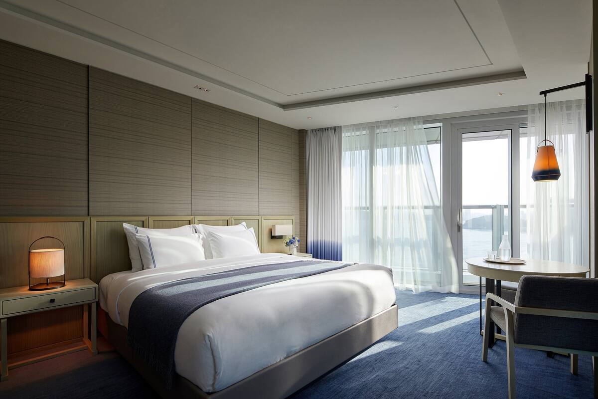 A hotel room with a large bed, a bedside table with a lamp, a chair, and a view of the outdoors through a large window.