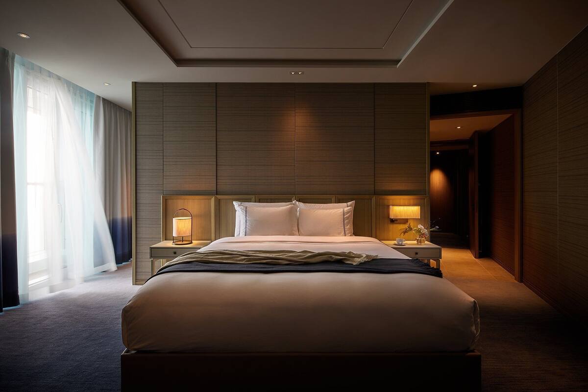 A modern hotel room with a large bed, two bedside tables with lamps, a large window with sheer curtains, and a wooden panel wall.