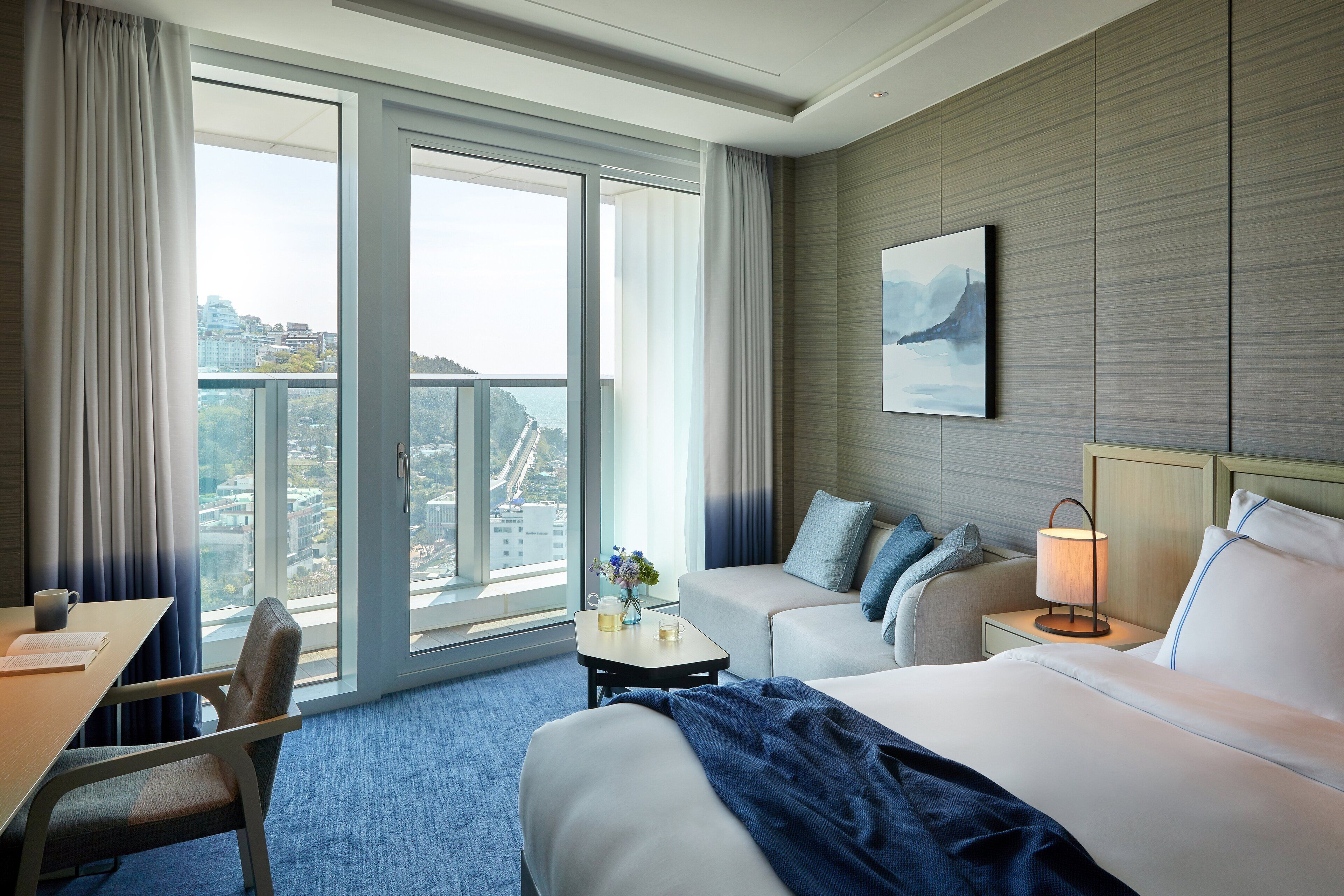 Et hotelværelse med stort vindue, seng, sofa, skrivebord og et natbord med lampe.