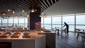 A modern dining area with a view of the sea, featuring a buffet with various food items and a person arranging tables. - Signiel Busan (Busan)