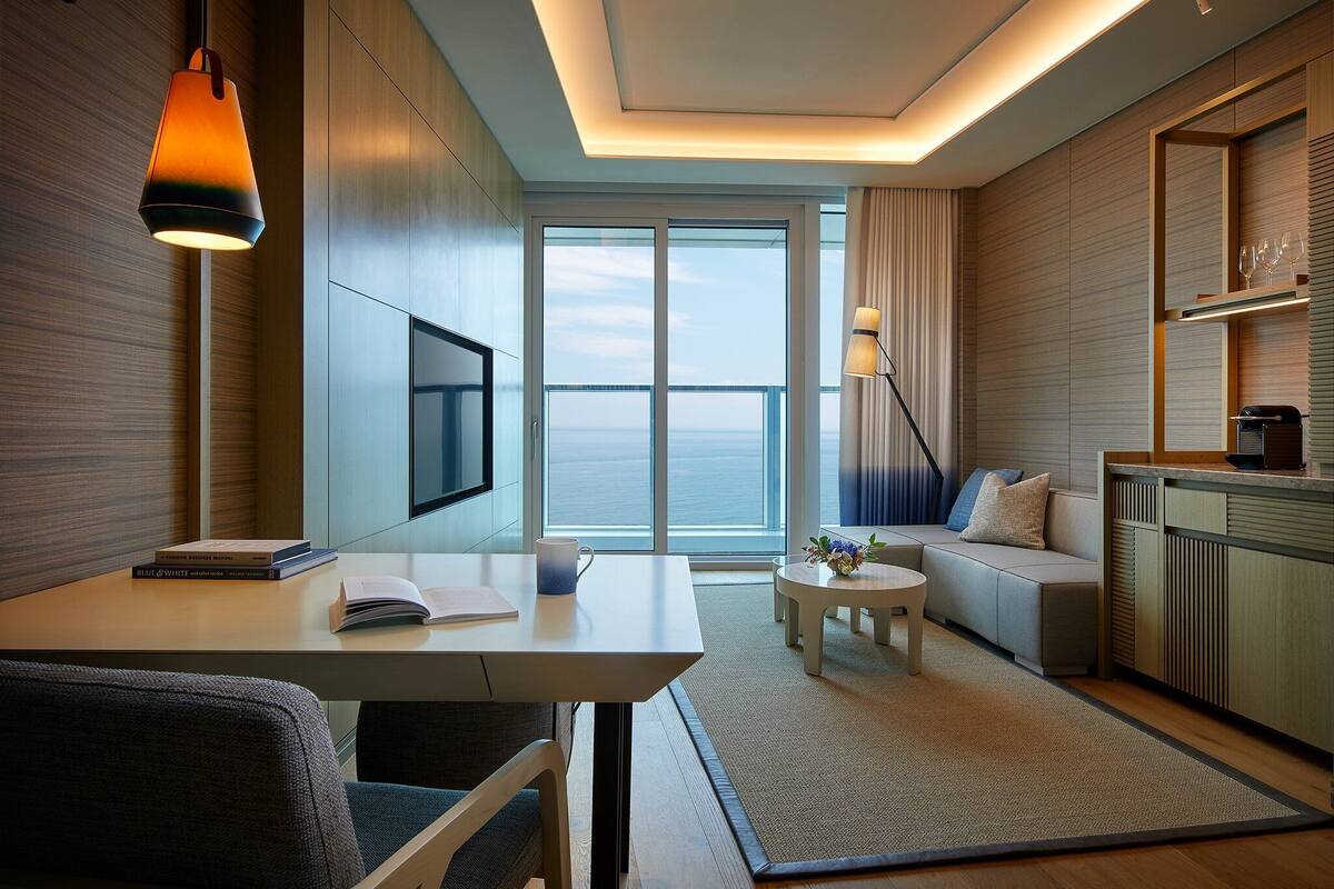 A modern hotel room with a large window, a flat-screen TV, a dining area, and a kitchen.