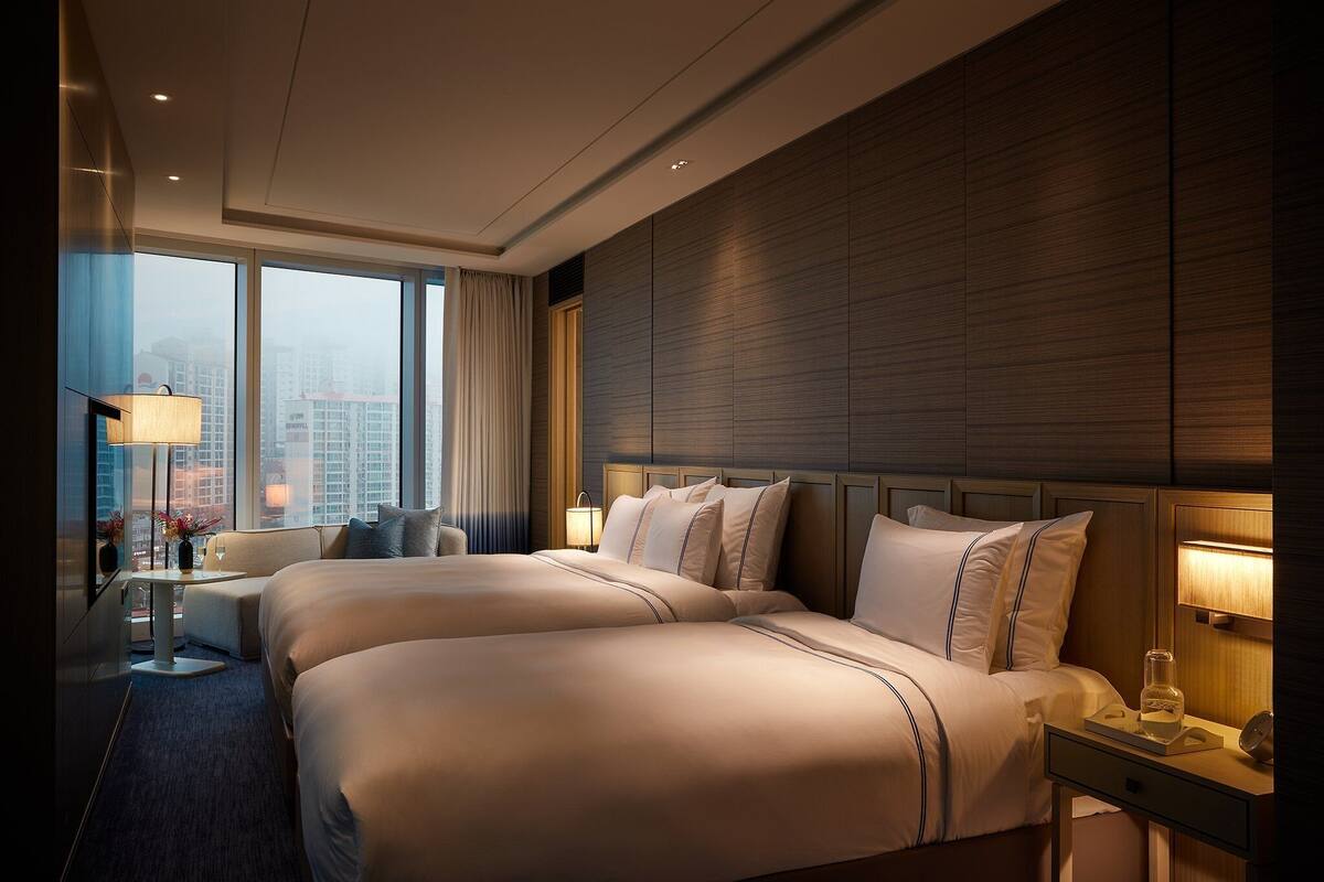 A hotel room with two beds, a desk, a lamp, and a view of the city.