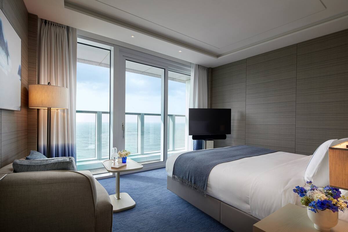 A modern hotel room with a large bed, a flat-screen TV, a desk with a lamp, a chair, and a view of the ocean through large windows.