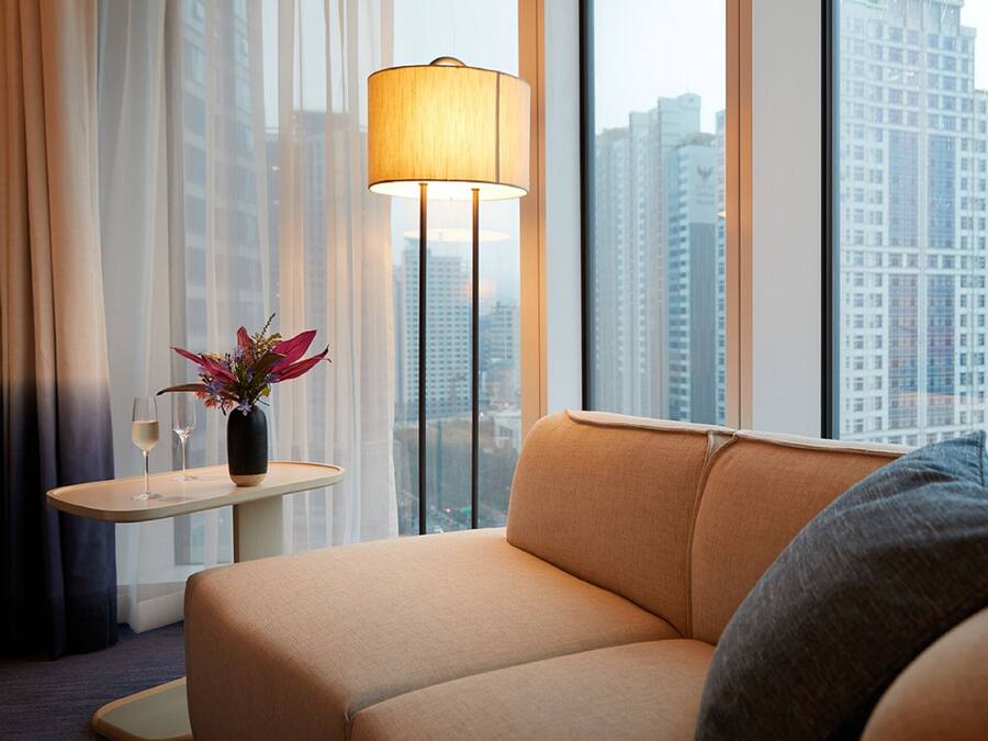A modern hotel room with a sofa, a small table with a lamp, and a view of tall buildings.