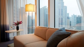 A modern hotel room with a sofa, a small table with a lamp, and a view of tall buildings.