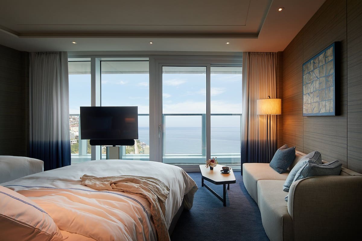 A hotel room with a large bed, a sofa, a small table, a TV, and a view of the sea through large windows.