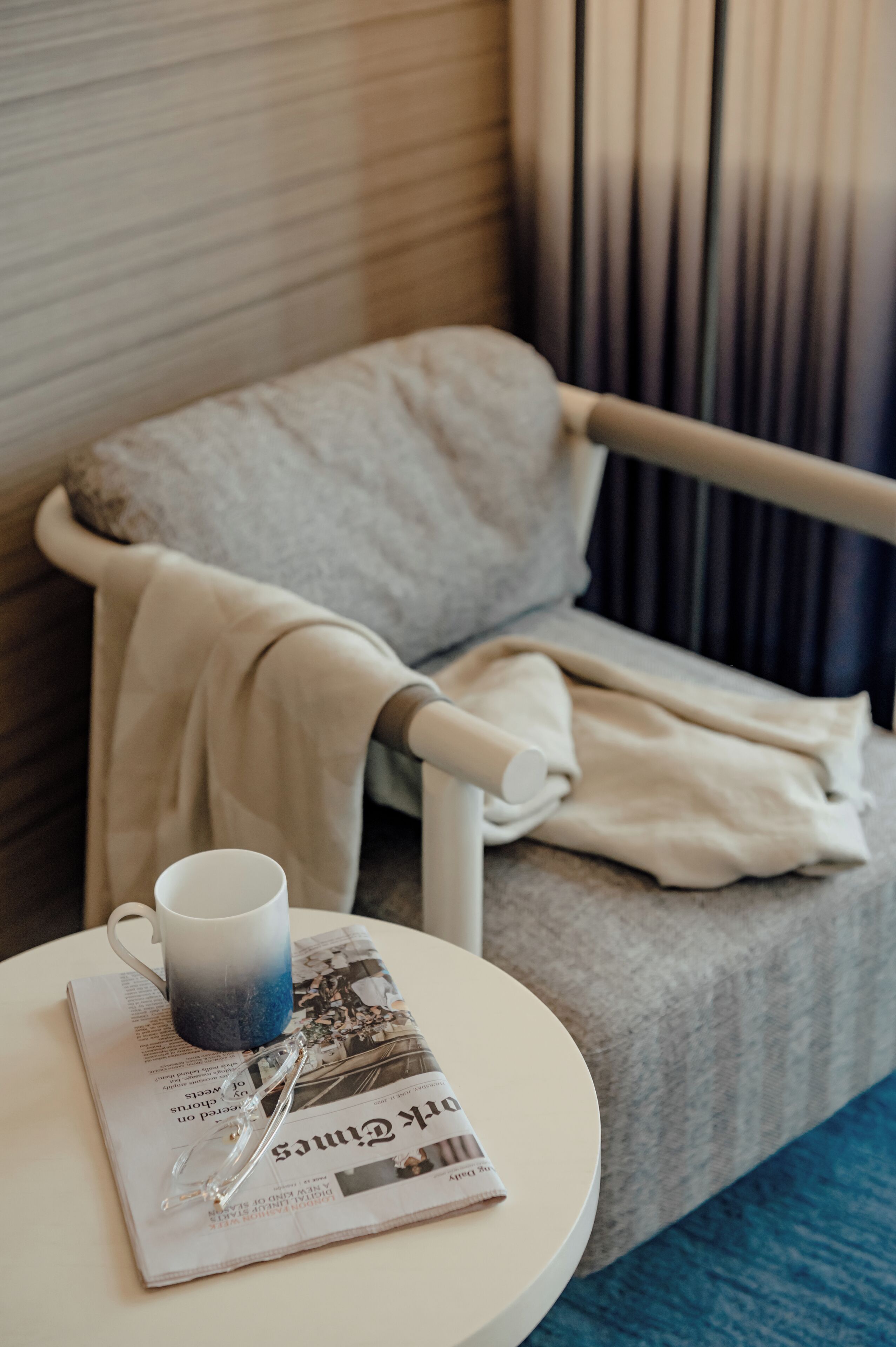 A neatly arranged hotel room with a bed, a bedside table, a newspaper, a mug, and a pair of glasses.