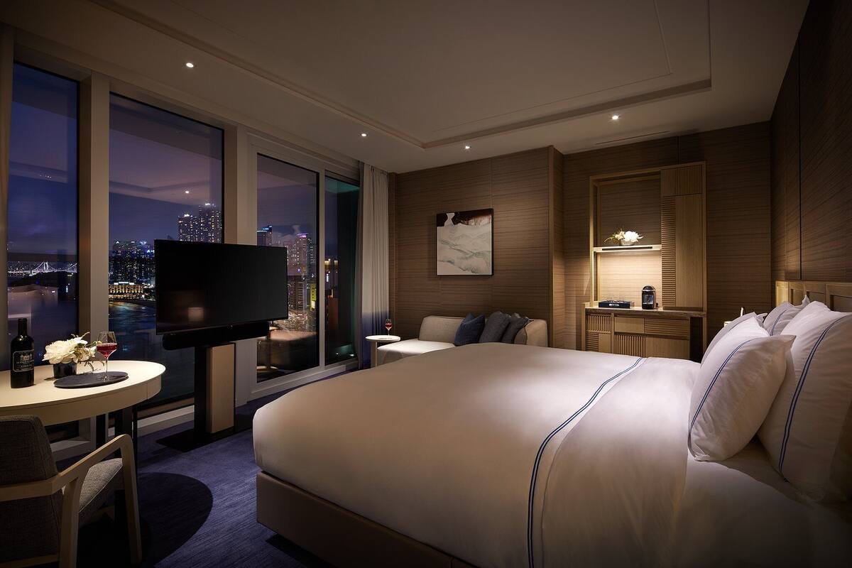 A hotel room with a large bed, a TV, a small table with a wine glass, and a view of the city at night.